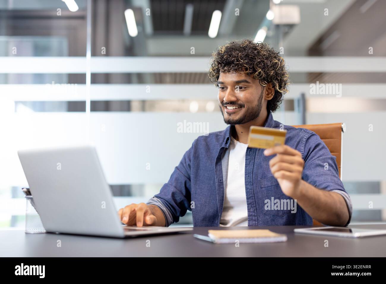 Sorridente giovane indiano che lavora in ufficio su un portatile e fa transazioni finanziarie con una carta di credito. Foto Stock