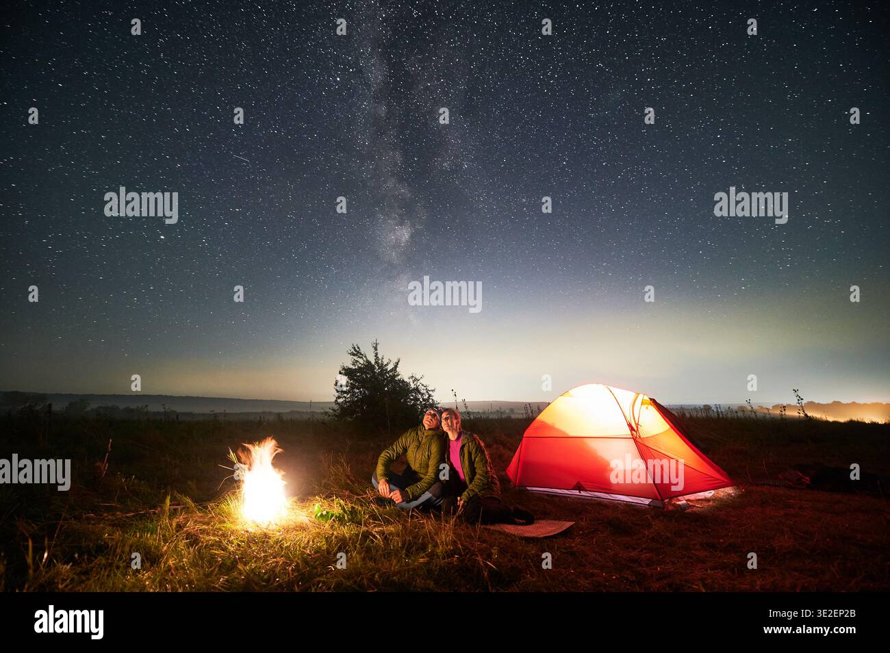 Splendida vista del cielo stellato notturno sul prato erboso con una coppia vicino al falò. Uomini e donne escursionisti seduti vicino al falò luminoso e guardando il cielo con le stelle. Concetto di trekking e campeggio notturno. Foto Stock