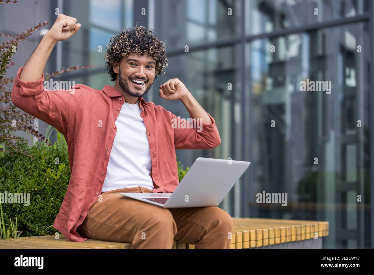 Ritratto di un uomo indiano felice in una camicia a righe seduto su una panchina all'esterno, tenendo un portatile in grembo, gioendo e alzando le braccia. Foto Stock