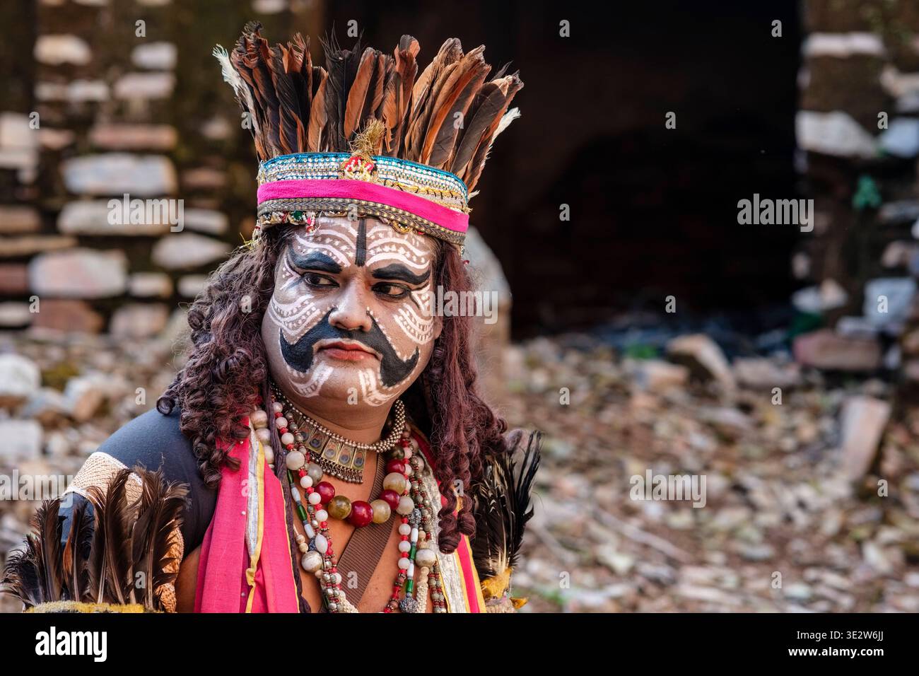 Un artista al festival Bundi Utsav indossa elaborati colori per il viso e un copricapo piumato di fronte a una porta di pietra scura. Bundi Rajasthan India F. Foto Stock