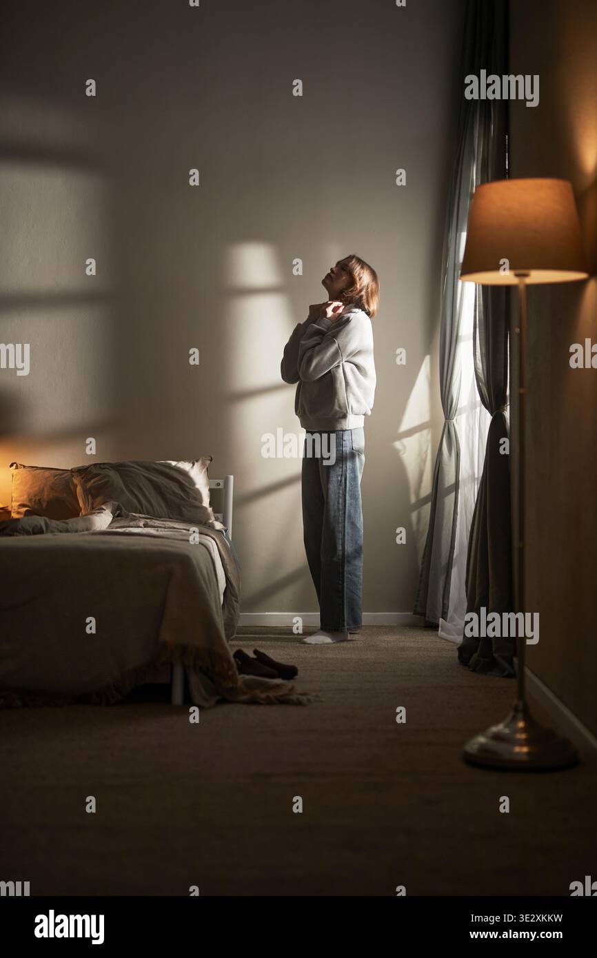 Verticale della giovane donna caucasica adulta in piedi vicino alla finestra in camera da letto, tenendo il collo con entrambe le mani mostrando espressione contemplativa o emotiva con ombre soffuse che proiettano luce Foto Stock