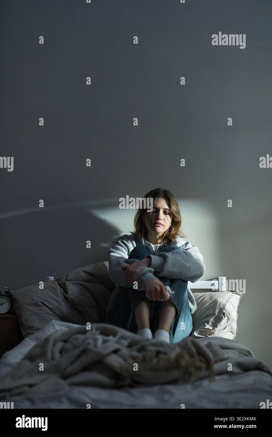 Verticale di una giovane donna caucasica seduta sul letto che abbraccia le ginocchia, guarda avanti con una triste espressione facciale, cuffie nelle orecchie, luce del mattino che proietta ombra sul muro, trasmettendo emozioni Foto Stock