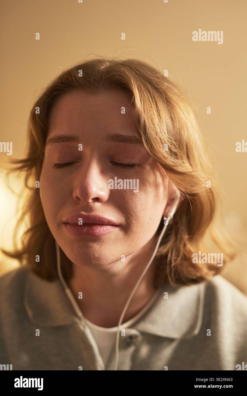 Verticale di una giovane donna caucasica adulta che chiude gli occhi con espressione emotiva, ascolta la musica attraverso auricolari cablati, mostra sottili lacrime sulle guance, trasmettendo sensazioni profonde Foto Stock