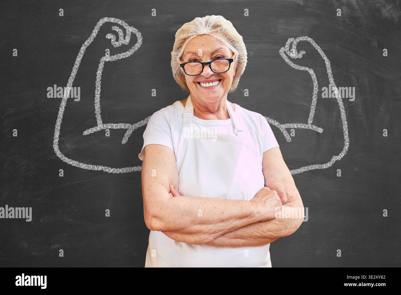 Felice donna anziana che cuoce davanti a un tavolo con braccia muscolari, che rappresenta la competenza. Foto Stock