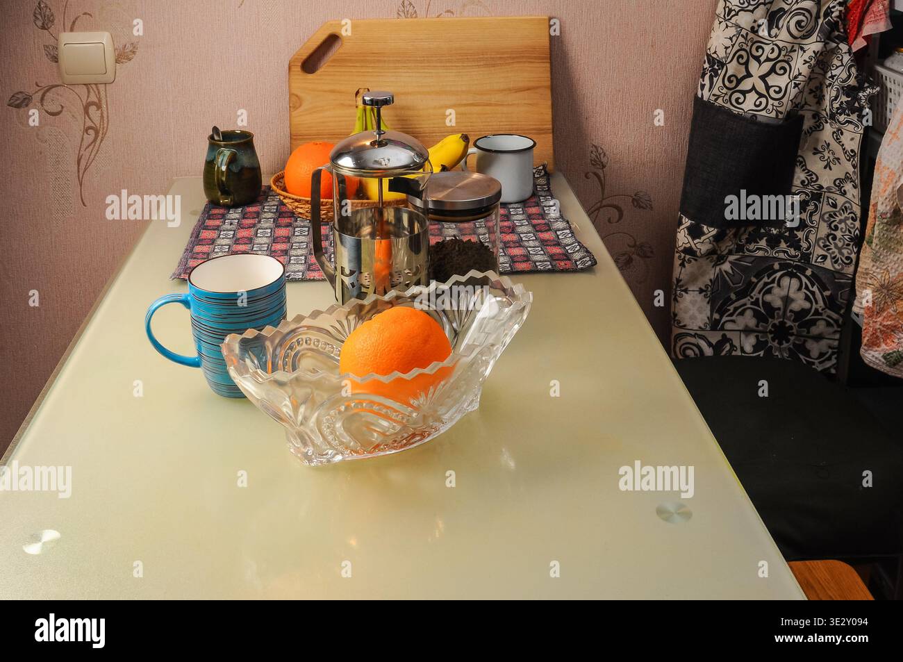 Una tazza blu brillante si trova accanto a una ciotola di cristallo che regge un'arancia succosa, con una pressa francese e banane sullo sfondo su un tovagliolo a motivi geometrici Foto Stock