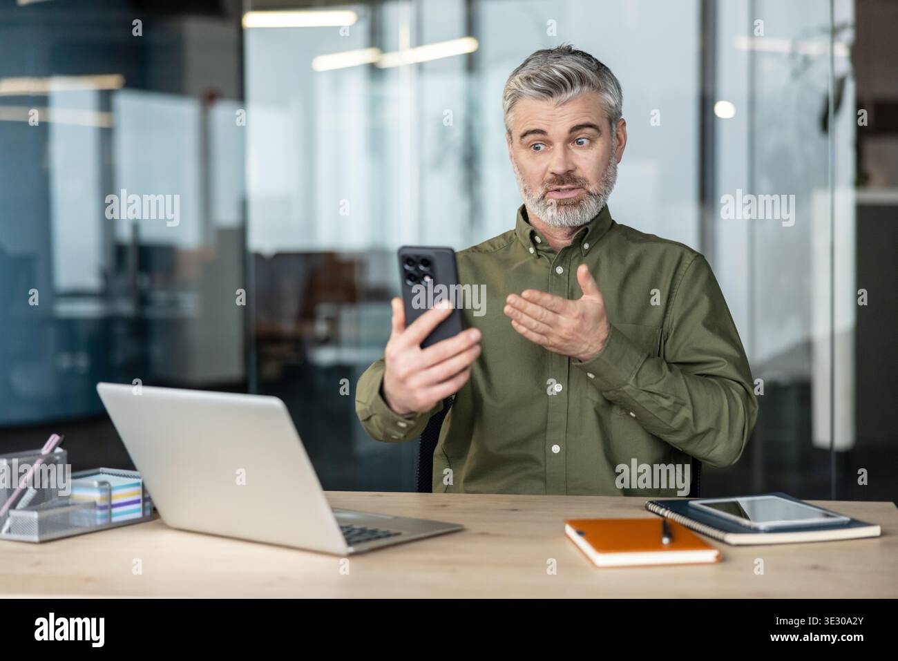 Uomo d'affari fiducioso dai capelli grigi che ha un'importante videochiamata su uno smartphone mentre è seduto alla sua scrivania in un ufficio moderno e comunica in remoto tramite la tecnologia Foto Stock