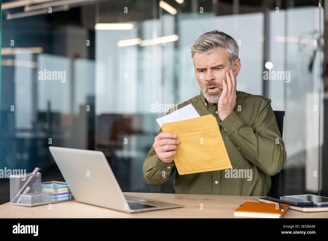 Un uomo maturo scioccato seduto a una scrivania, che apre una busta importante con notizie inaspettate o un conto di grandi dimensioni, che subisce lo stress finanziario in un ufficio moderno Foto Stock