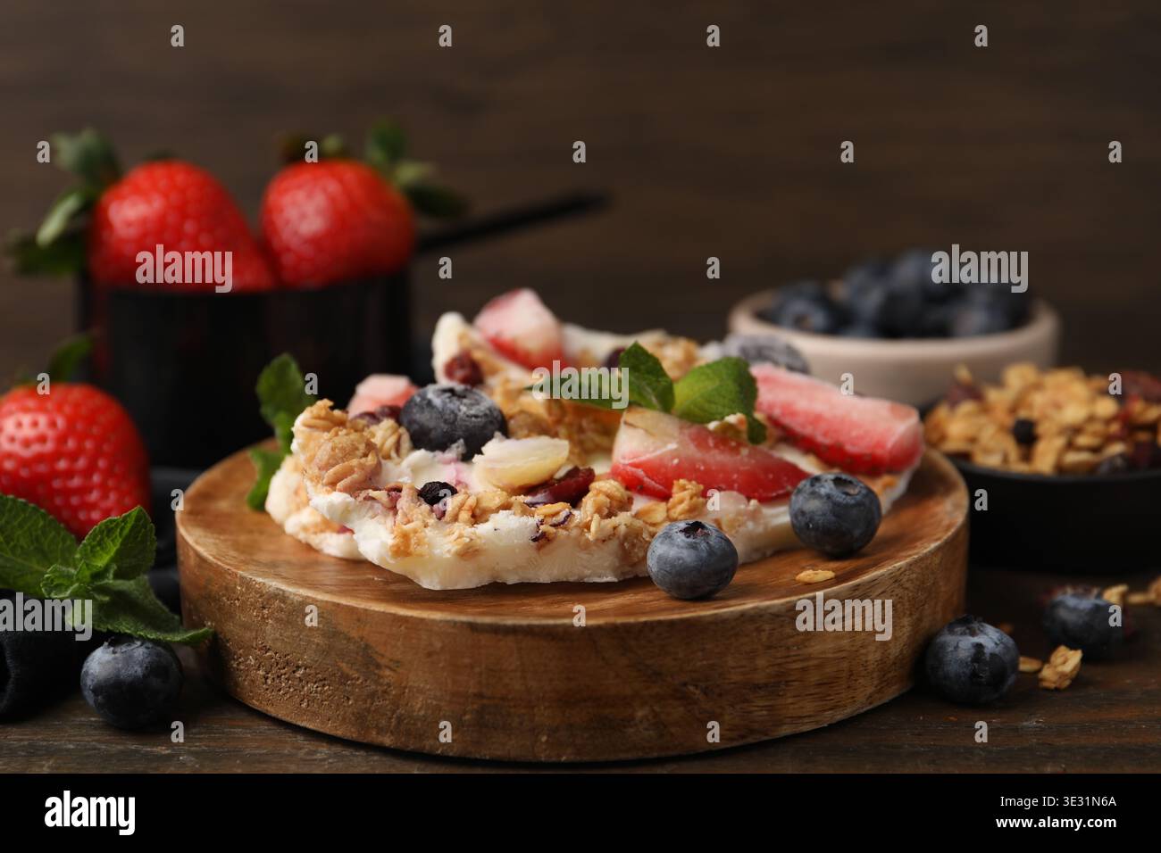 Pezzi di gustosa corteccia di yogurt con frutti di bosco, menta e muesli su tavola di legno, primo piano Foto Stock