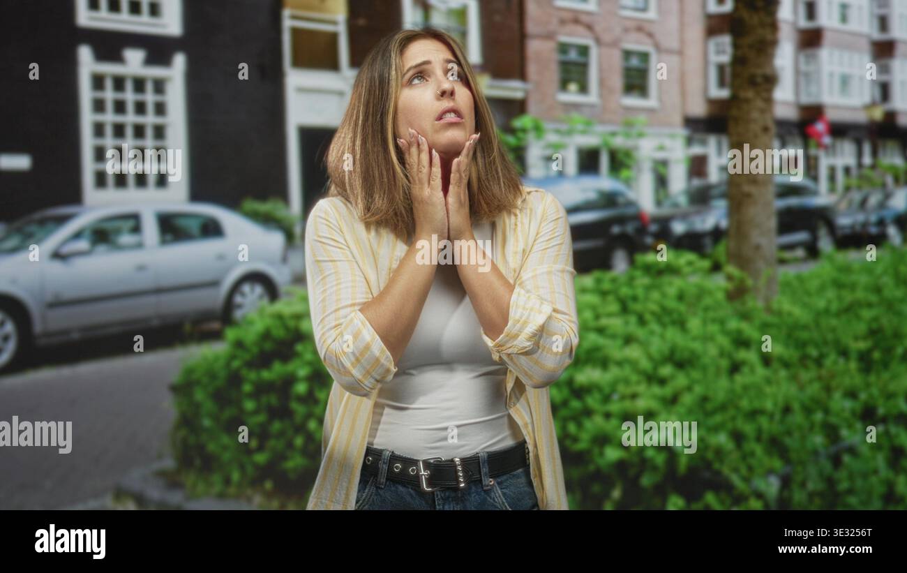 Donna che preme le palme alle guance con un'espressione dolorosa su una strada della città, accanto a auto parcheggiate e siepe verde davanti a case a schiera; preoccupati. Foto Stock