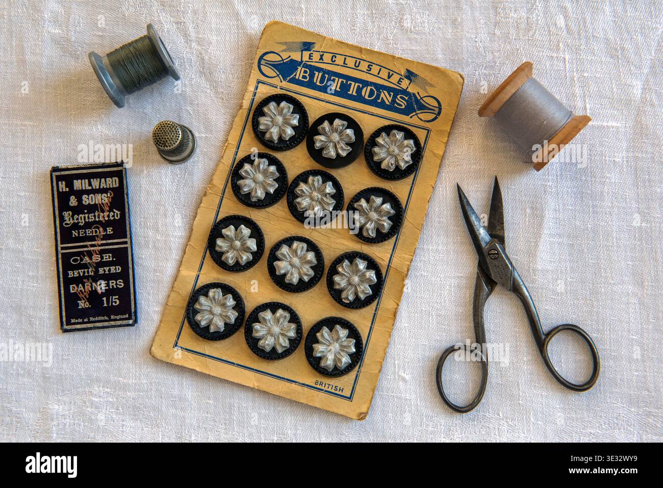 Kit pulsanti per cucire. Tessera di bottoni vintage neri e argentati con pacco di aghi, bobine di cotone grigio, forbici e ditale su panno bianco. Foto Stock