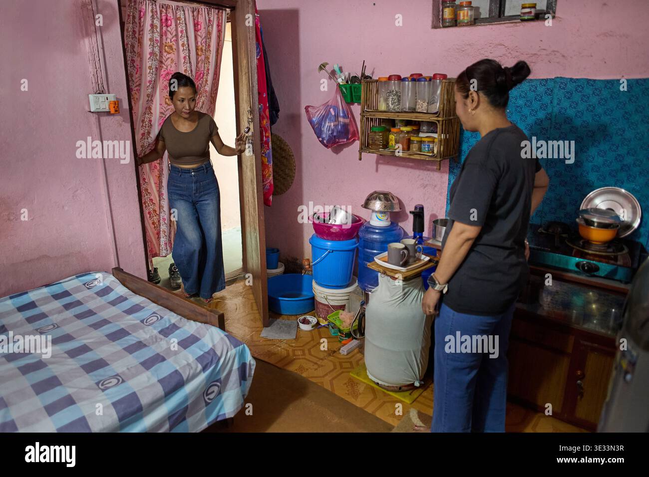 Uno sguardo alla vita quotidiana: Due donne che interagiscono in una stanza modesta con vivaci pareti rosa e semplici articoli per la casa Foto Stock