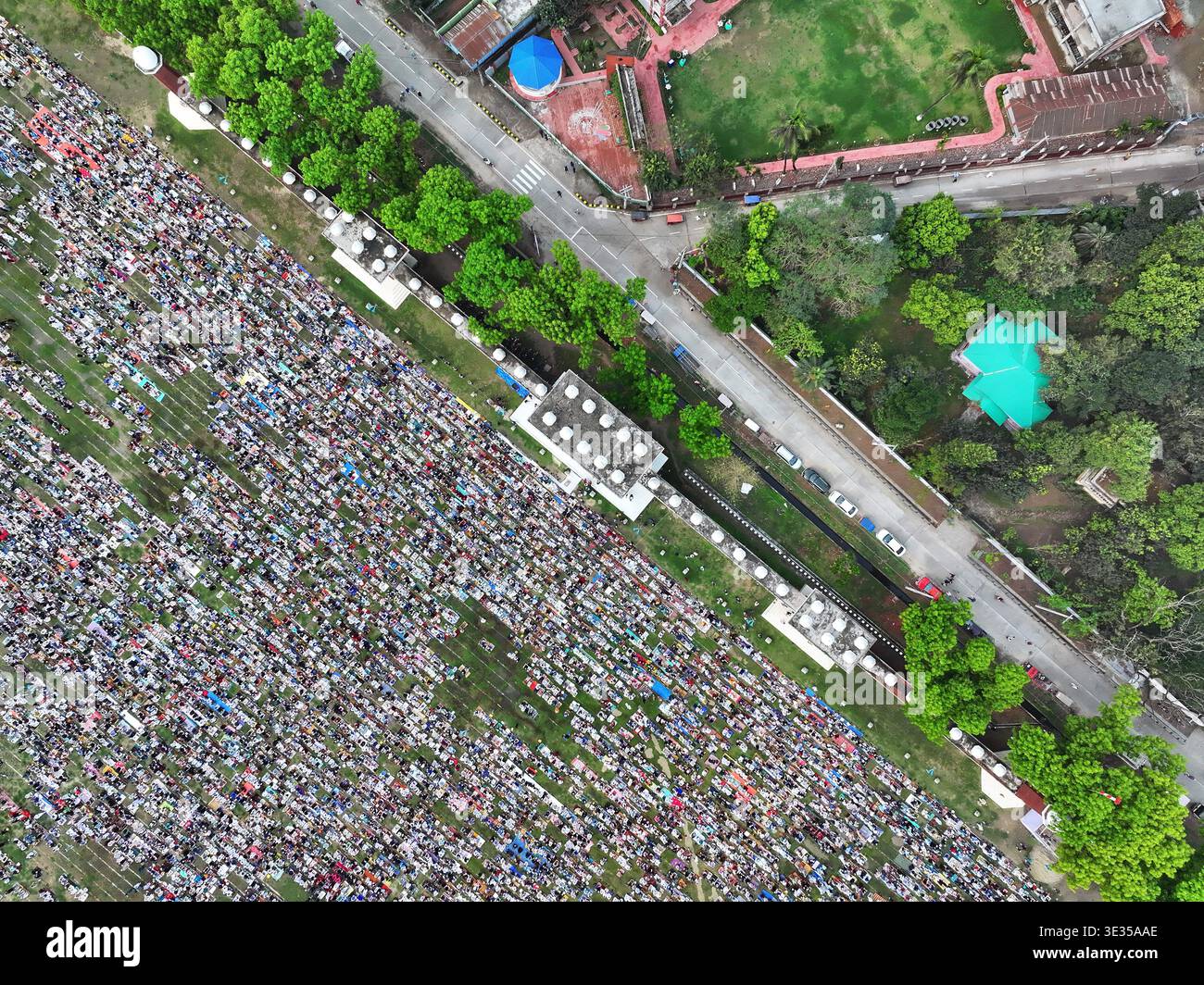 Dinajpur, Bangladesh - 21 marzo 2026: Veduta aerea di un mare di persone riunite su un vasto campo verde vicino a una strada costeggiata da alberi, adiacente alla costruzione Foto Stock