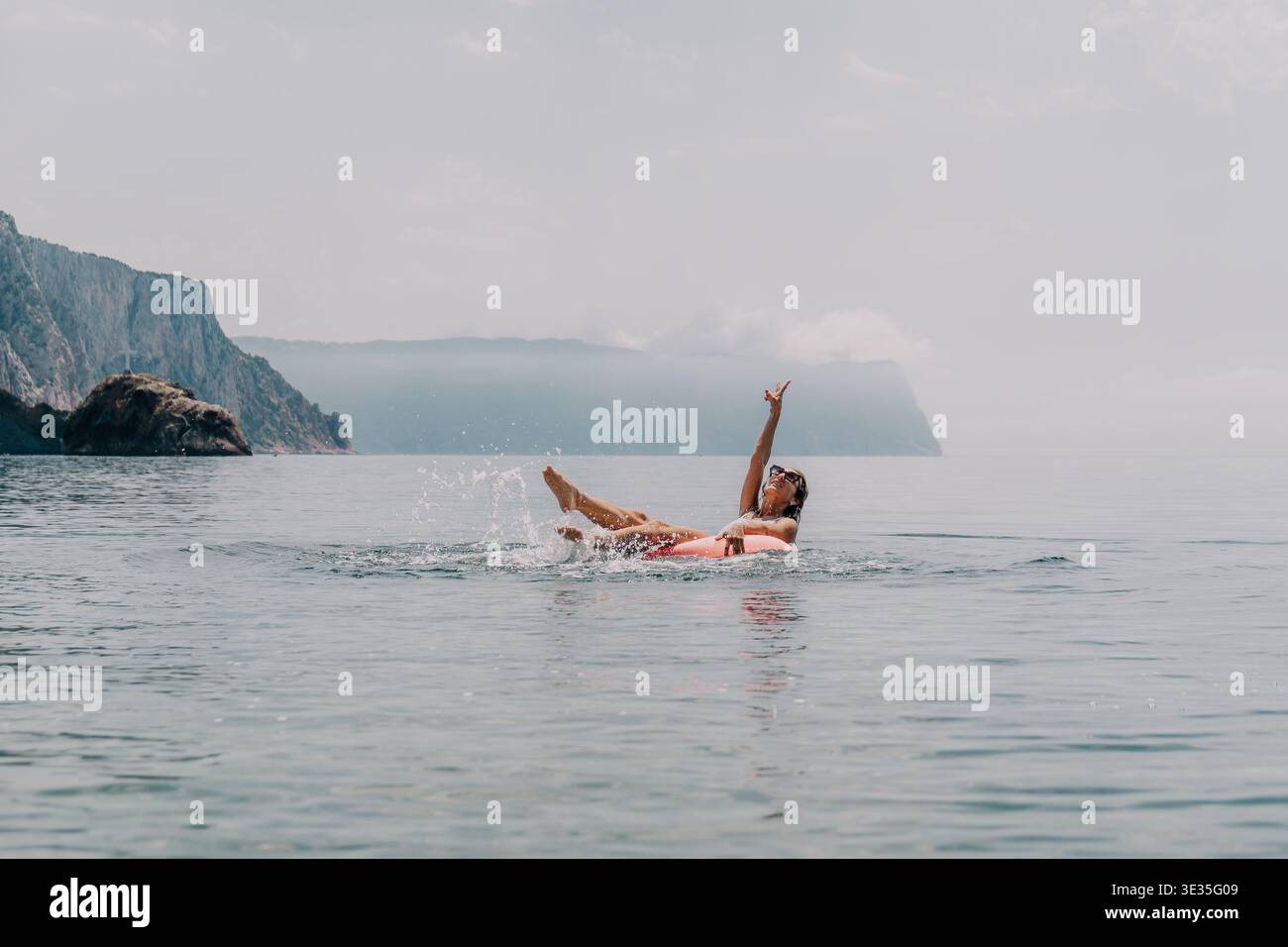 Donna che nuota spruzza giovane donna che galleggia su un anello gonfiabile in mare facendo spruzzi d'acqua con montagne panoramiche alle spalle Foto Stock