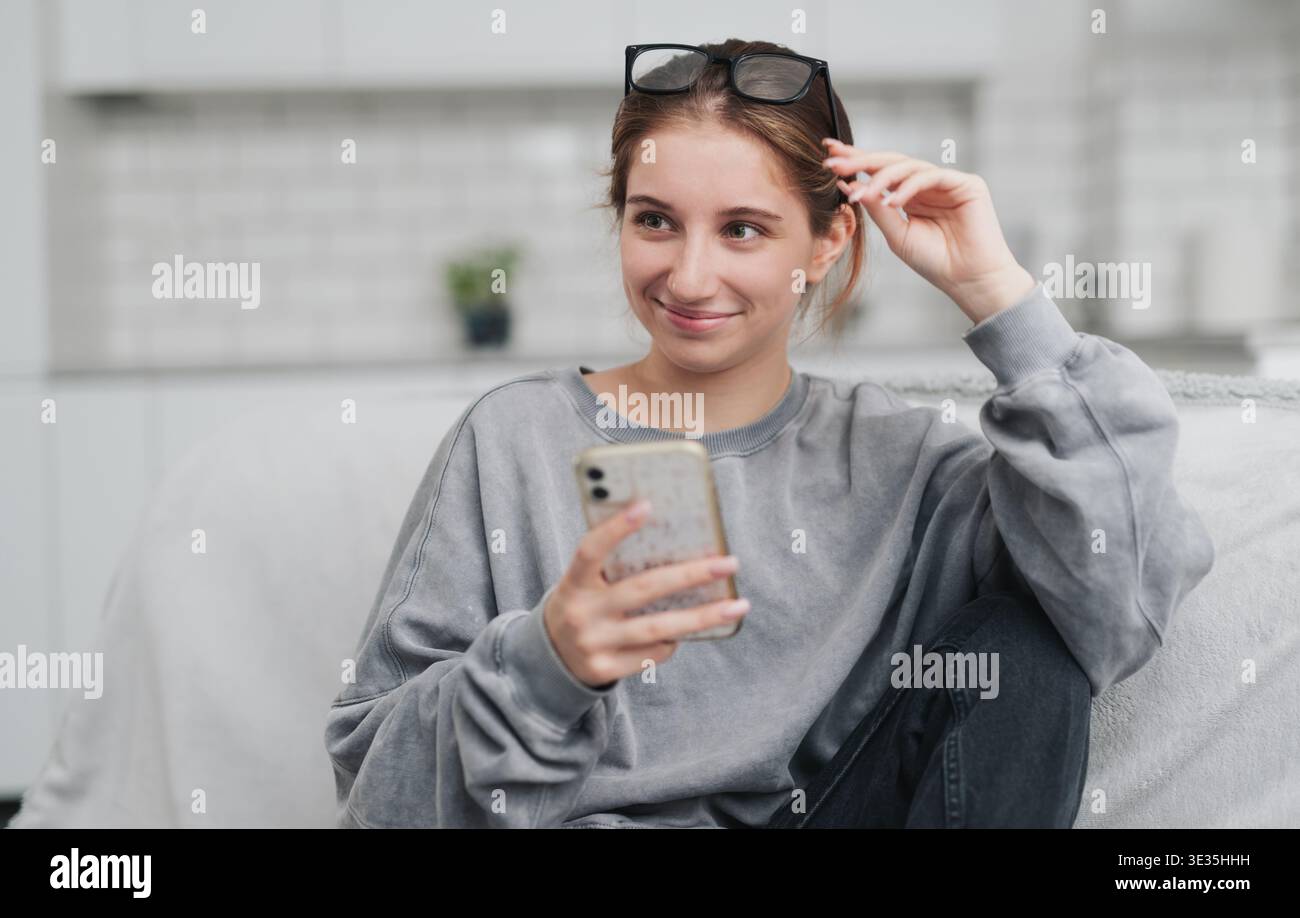Pretty Girl seduta su Un divano in Una stanza luminosa, che naviga su Internet con uno smartphone Foto Stock