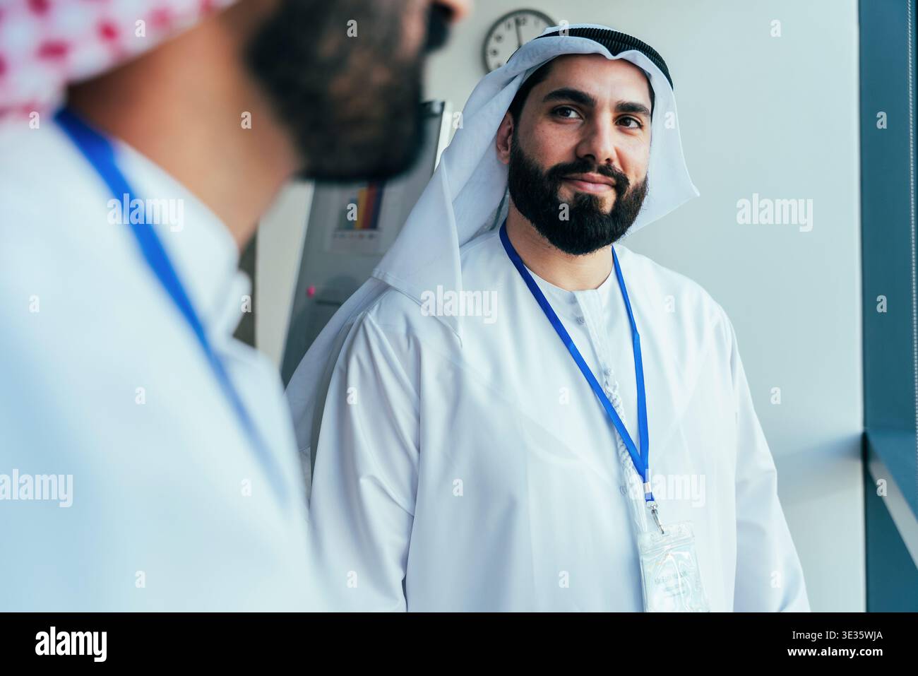 Uomo del Medio Oriente che indossa kandura e ghutra tradizionali, con barba e badge d'identità, ascolta una conversazione con un collega in un'azienda Foto Stock