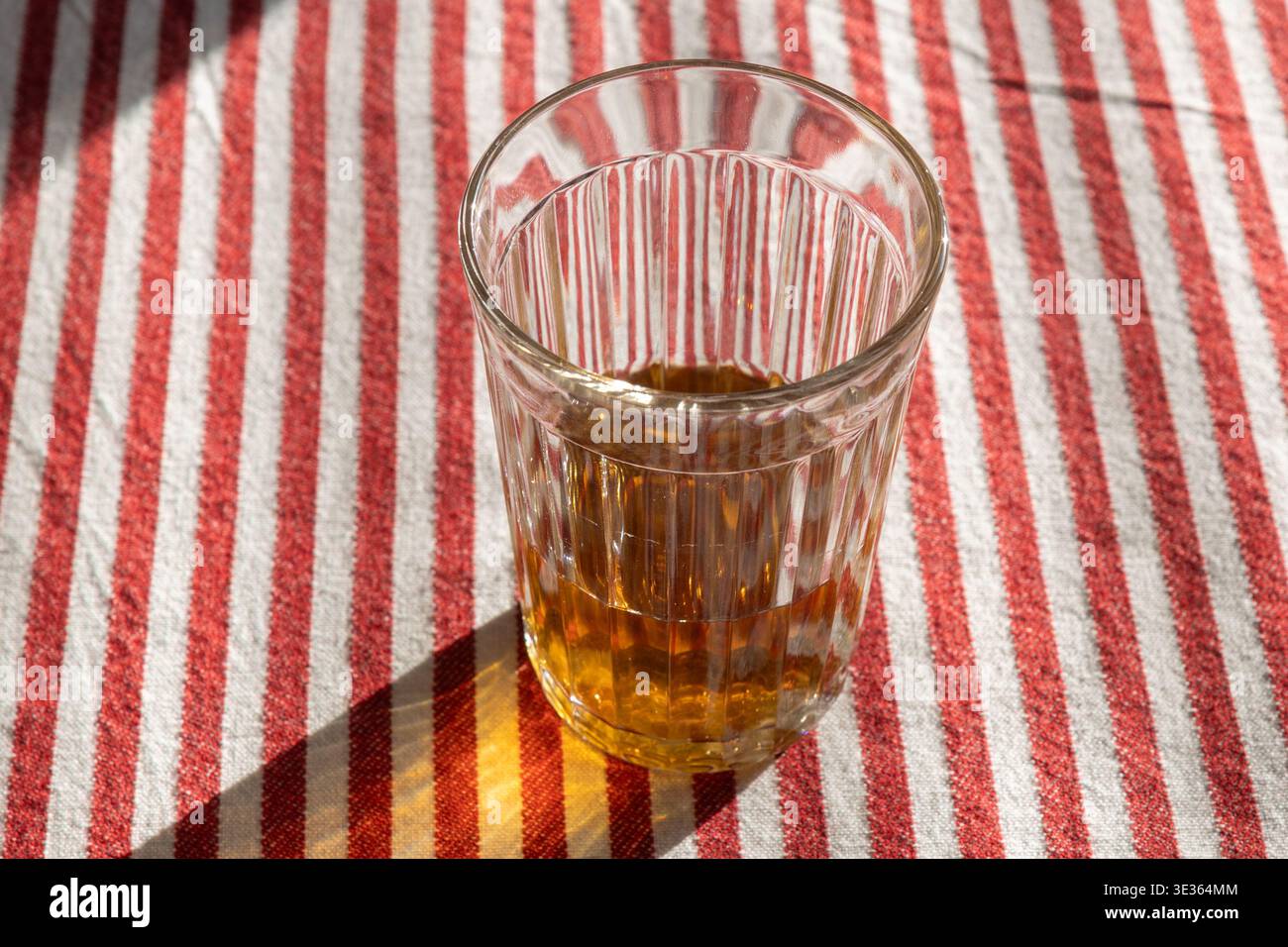Un bicchiere di tè siede su una tovaglia a strisce rosse e bianche, catturando la luce del sole in un ambiente luminoso e naturale Foto Stock
