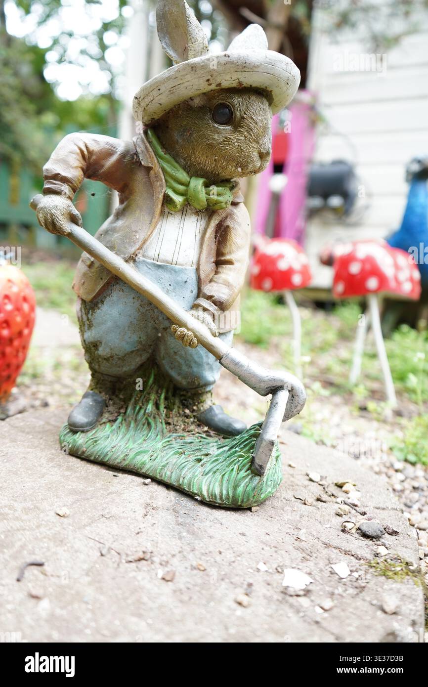 Stravagante ornamento da giardino di un coniglio vestito con un gilet e un cappello, che regge una zappa da giardino, ambientato in un giardino delle fate in miniatura Foto Stock