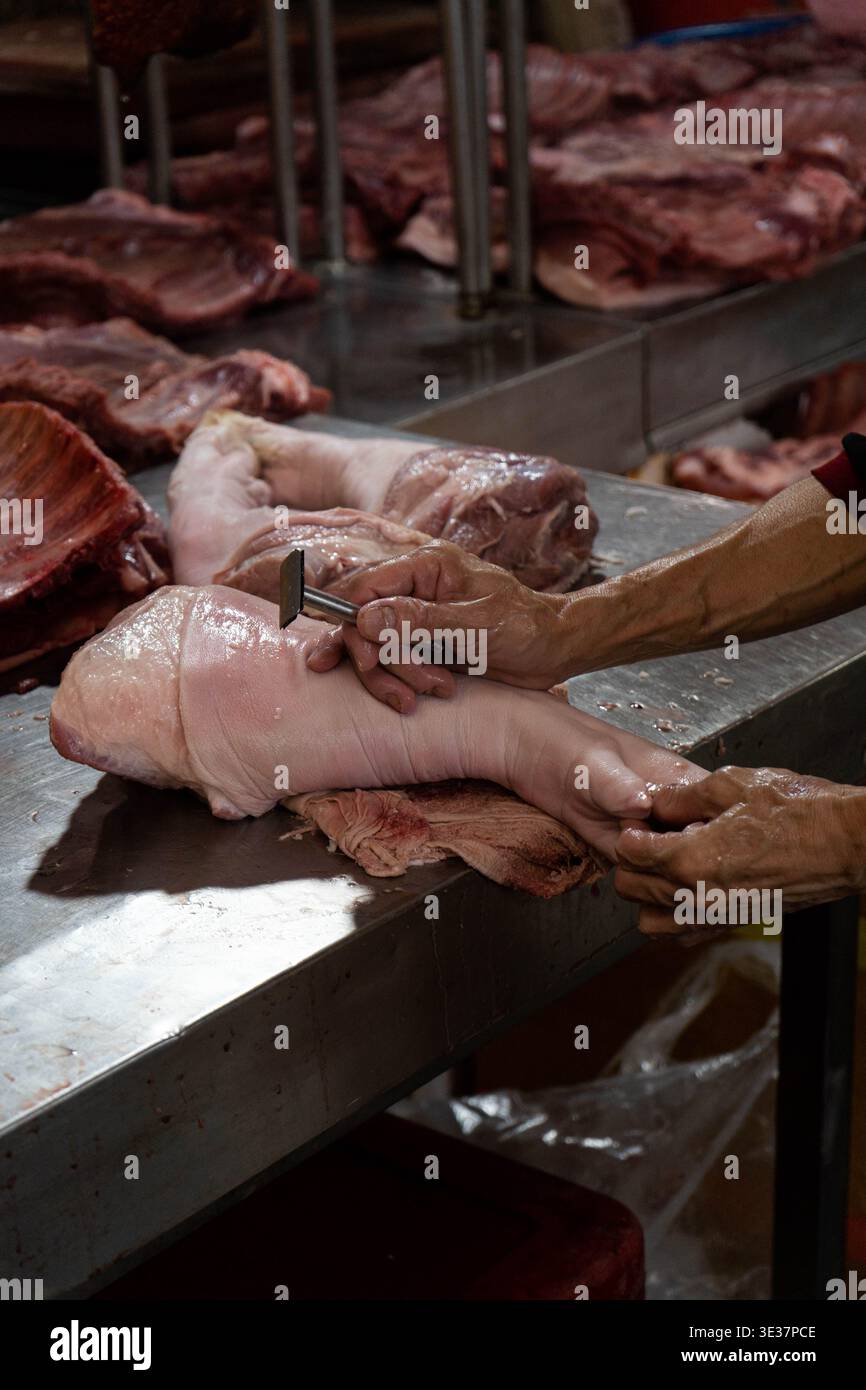 Un macellaio prepara una coscia di maiale in una bancarella del mercato locale, mostrando la lavorazione tradizionale della carne e l'attività quotidiana del mercato. Foto Stock