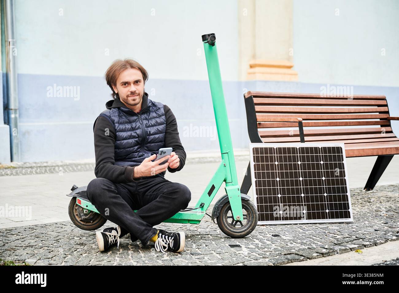 Ritratto della persona maschile seduto su uno scooter elettrico con pannello solare e smartphone durante la ricarica. Integrazione delle energie rinnovabili nel trasporto personale, evidenziando uno stile di vita urbano sostenibile. Foto Stock