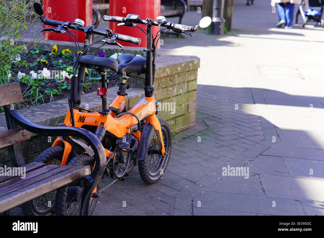 Bicicletta elettrica pieghevole arancione parcheggiata su una panchina di legno nelle soleggiate strade di Lyndhurst, New Forest, Hampshire, Inghilterra. Lyndhurst, Inghilterra Foto Stock
