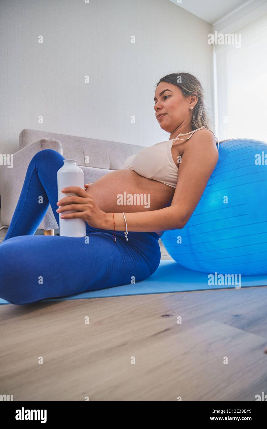 Donna incinta stanca che beve acqua dopo la sua sessione di allenamento su un tappetino da yoga in salotto Foto Stock