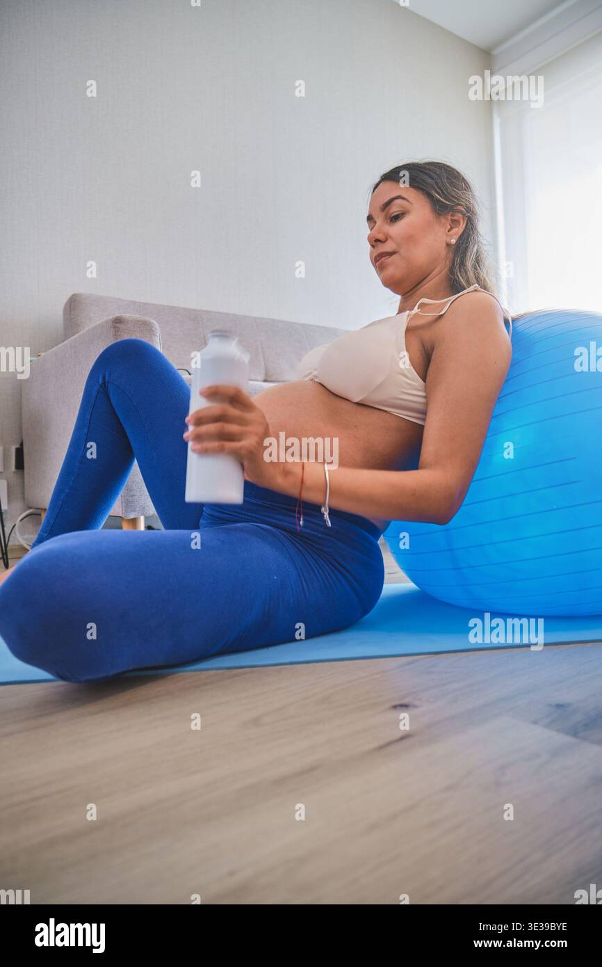Donna incinta che riposa su un tappetino da yoga con una palla da ginnastica e in mano una bottiglia d'acqua Foto Stock