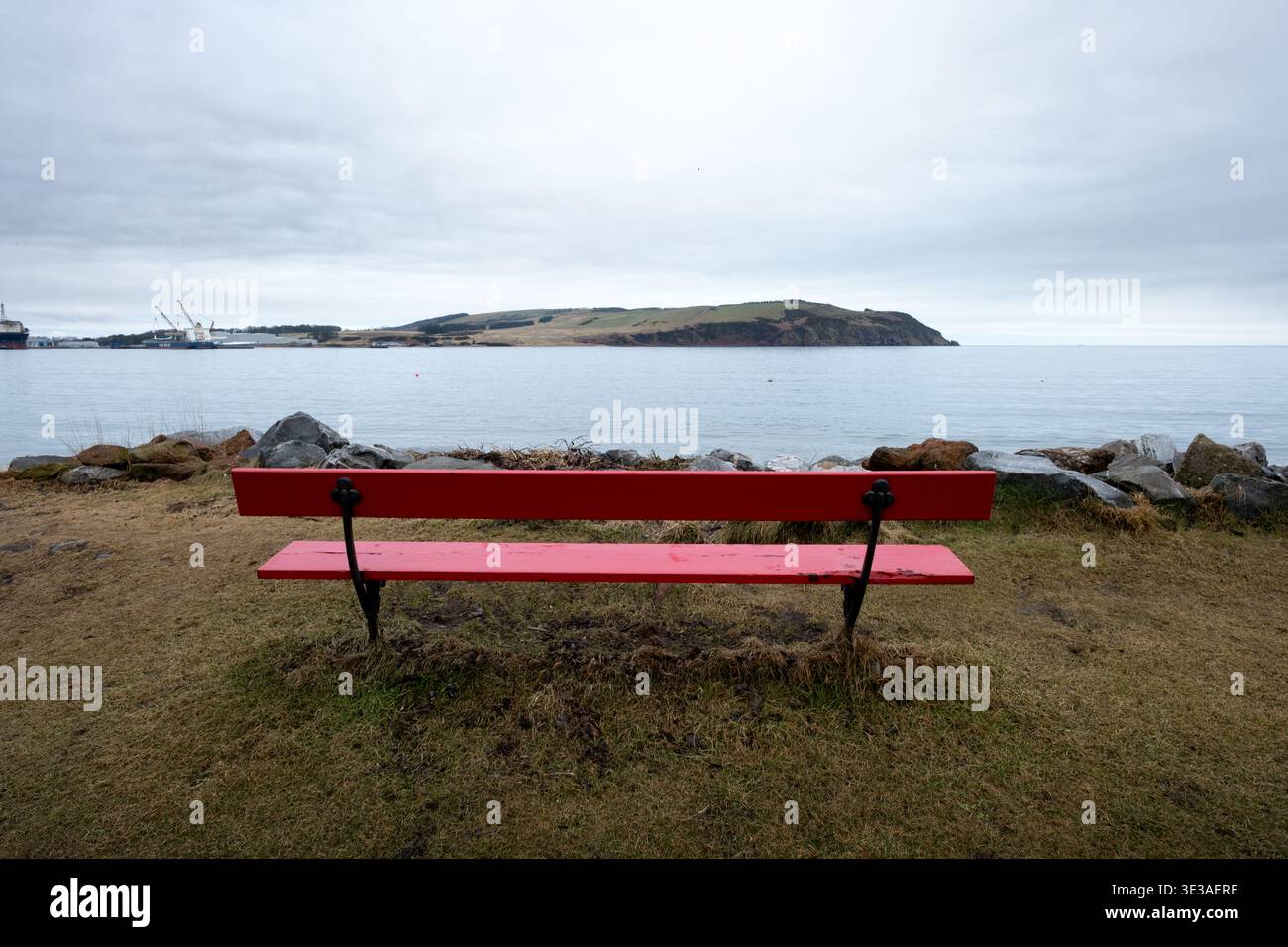 Panchina rossa con vista su Cromarty Firth a Cromarty, Highland, Scozia, con acque calme, costa rocciosa e cielo coperto. Foto Stock