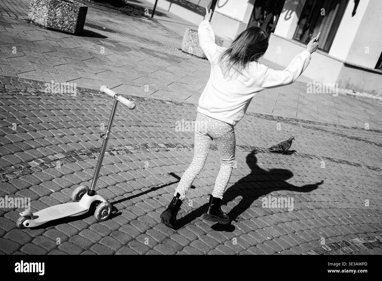 Il bambino si diverte e insegue il piccione sulla piazza della città con lo scooter. Momenti divertenti di tutti i giorni in un ambiente urbano in bianco e nero. Foto Stock