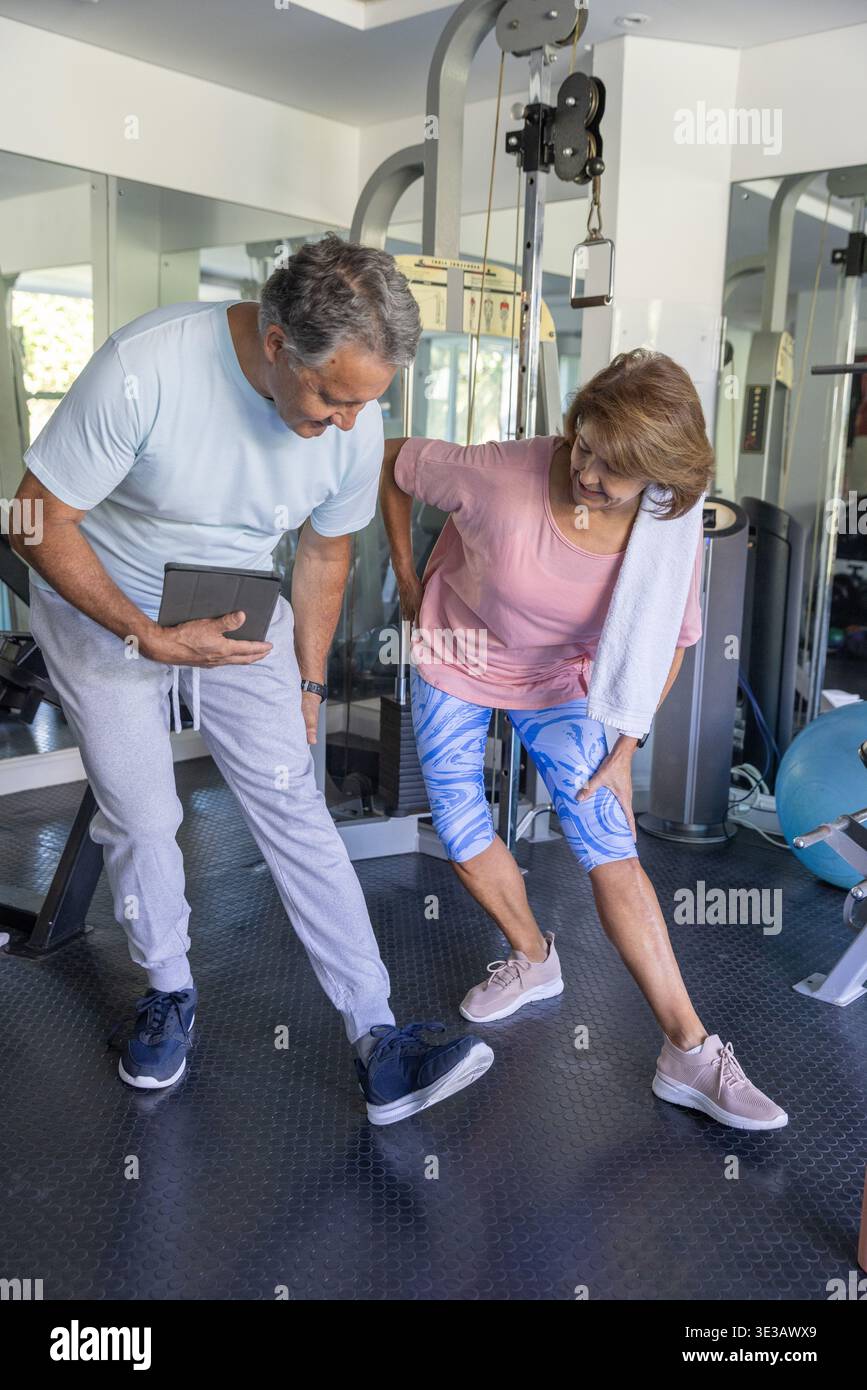 Donna anziana e uomo che si allenano in una palestra privata, tenendo un tablet e un asciugamano, specchi Foto Stock