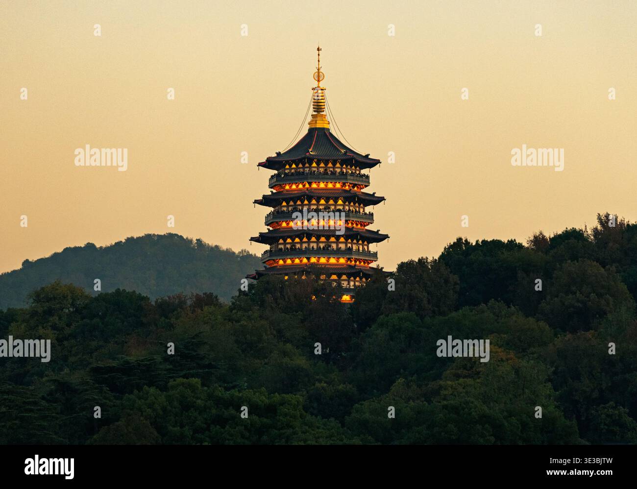 Tradizionale pagoda dell'Asia Orientale che brilla di calde luci al tramonto, annidata tra lussureggianti alberi verdi su una tranquilla collina Foto Stock