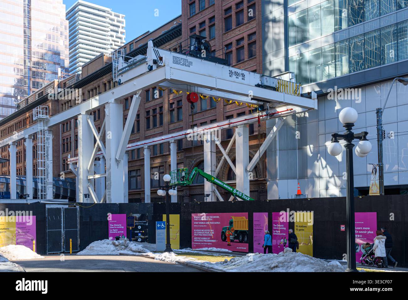 La grande gru a cavalletto bianca si estende su un cantiere del centro tra gli edifici, con la neve a terra e i pedoni che camminano nelle vicinanze. Foto Stock
