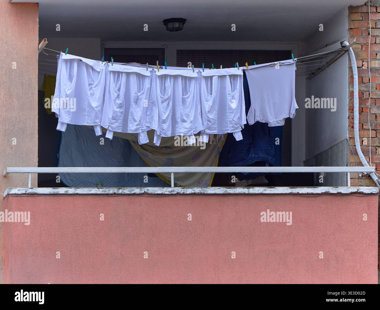 Camicie bianche appese su una linea di abbigliamento su un balcone con parete rosa e sfondo in mattoni, che mostrano la lavanderia quotidiana in un ambiente residenziale urbano Foto Stock