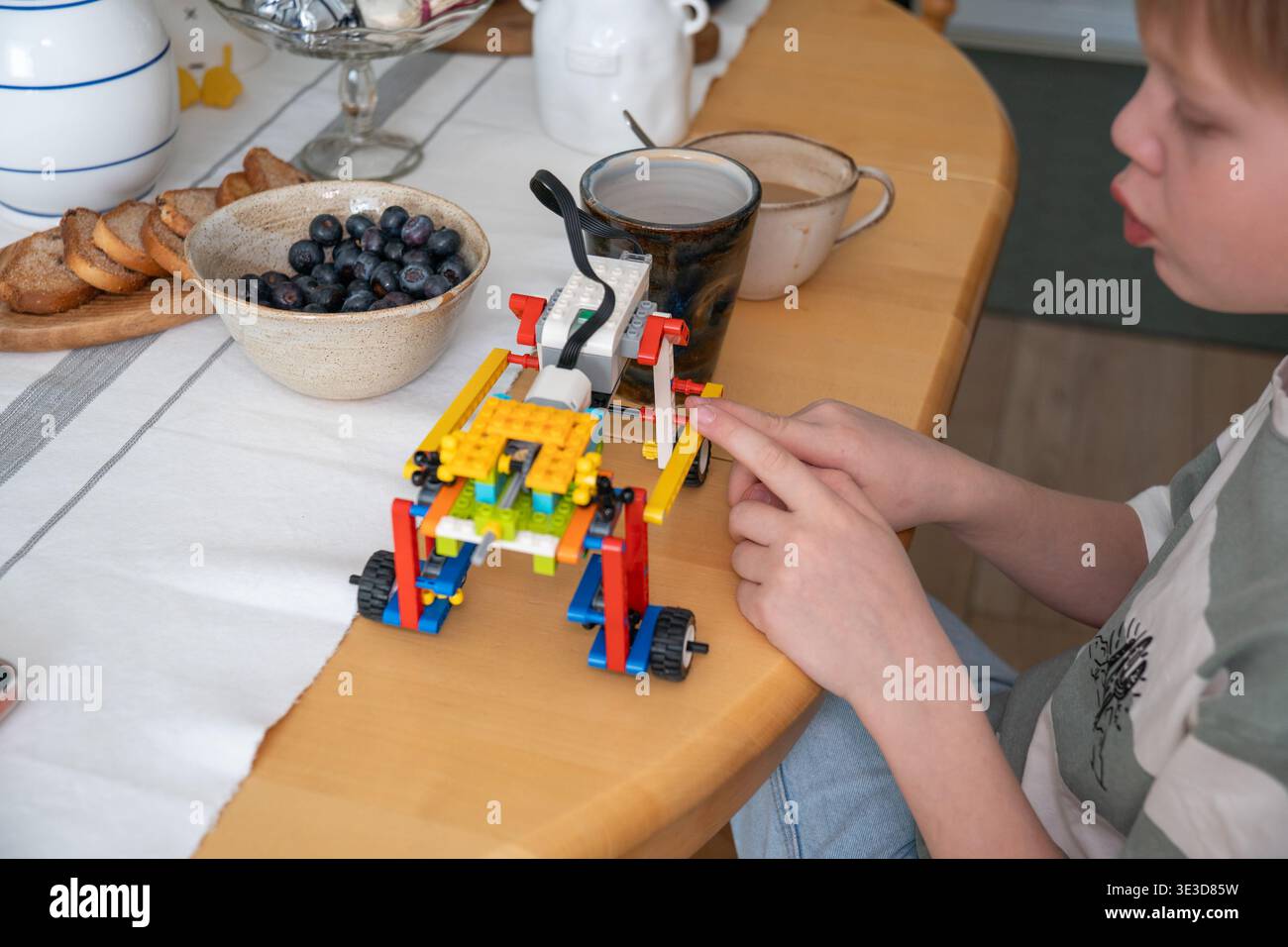 Il bambino assembla un giocattolo da costruzione a tavola con articoli da colazione in interni di casa con luce naturale, attività focalizzata e momenti di apprendimento quotidiani Foto Stock