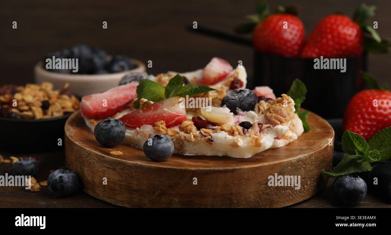 Pezzi di gustosa corteccia di yogurt con frutti di bosco, menta e muesli su tavola di legno, primo piano Foto Stock