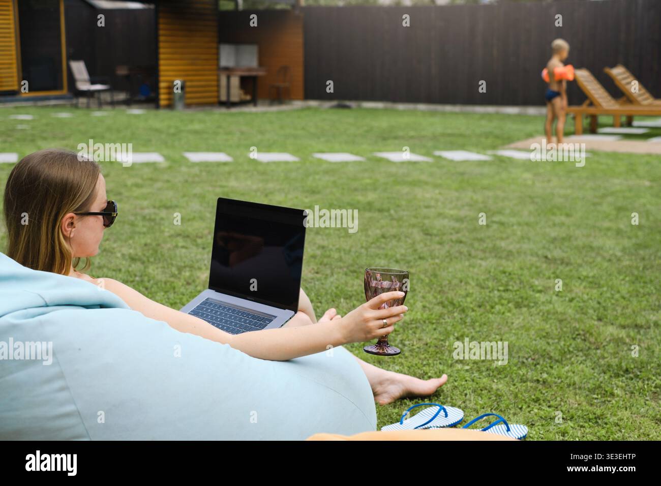 La donna freelance lavora con un computer portatile nel giardino del cortile con un bicchiere di vino mentre il figlio gioca in sottofondo. Concetto di lavoro a distanza e estate rilassante Foto Stock
