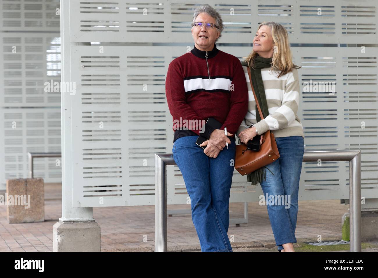 Coppia senior in piedi e appoggiata alle ringhiere di metallo del plaza, indossando maglioni e tenendo la borsa Foto Stock