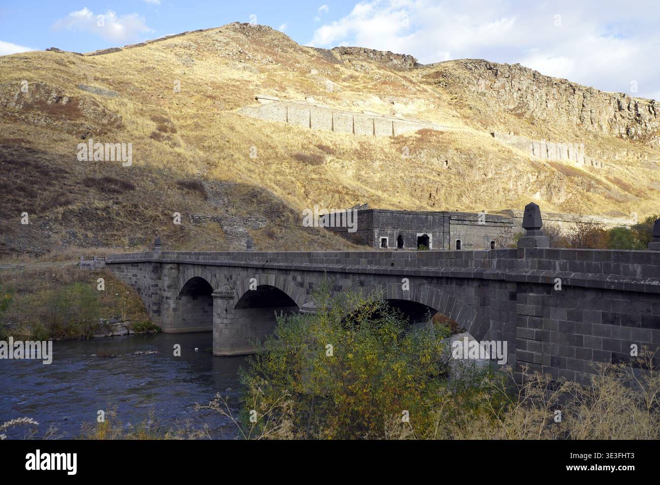 Un ponte ad arco in pietra di epoca russa attraversa il fiume Kars sotto un'aspra collina con fortificazioni storiche e rovine vicino a Kars, Turchia Foto Stock