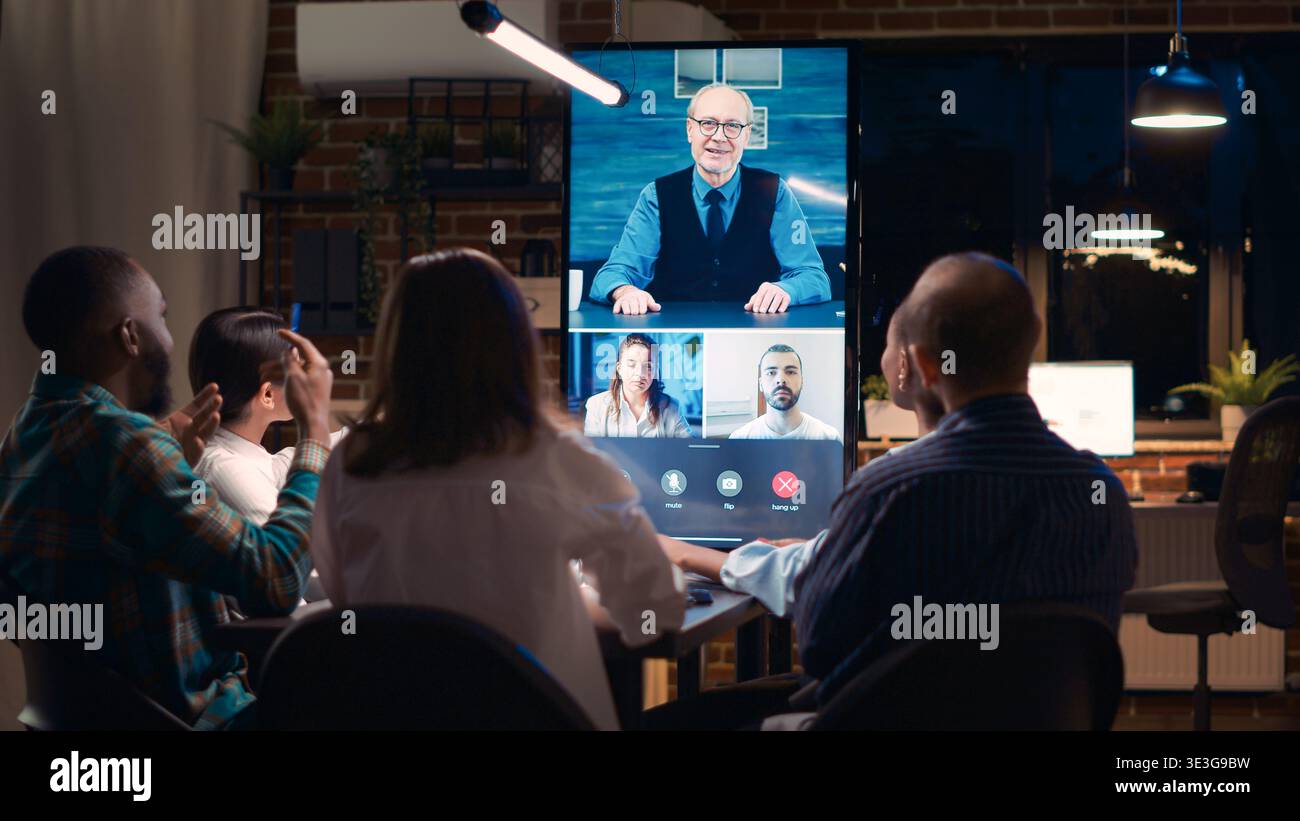 I dipendenti salutano i dirigenti anziani in videoconferenza, salutano i colleghi, comunicano in videoconferenza. Gli impiegati che ascoltano il responsabile del team che parla, che parla in teleconferenza di notte. Scatto con cavalletto. Foto Stock