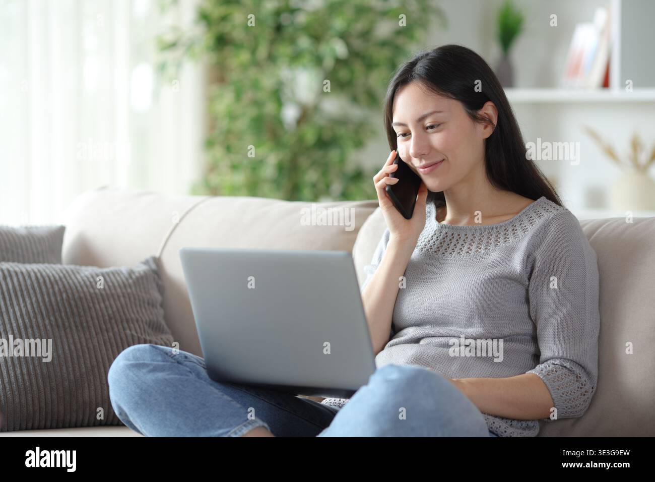 Donna asiatica soddisfatta che fa una telefonata sui contenuti online seduti sul divano di casa Foto Stock