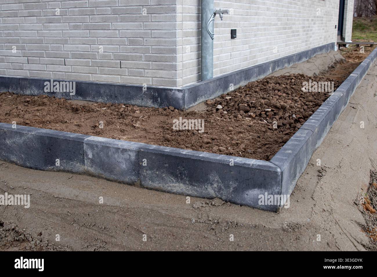 Bordatura in cemento installata intorno a un letto da giardino con terreno appena trasformato e una parete in mattoni di una casa visibile sullo sfondo in una giornata limpida Foto Stock