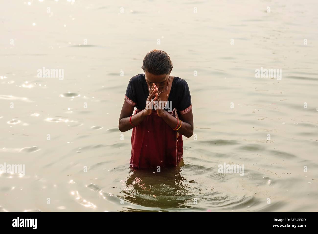 Una donna esegue una preghiera rituale mentre si trova nel fiume Gange a Varanasi, con la luce del sole che riflette sulla superficie increspata dell'acqua. Foto Stock