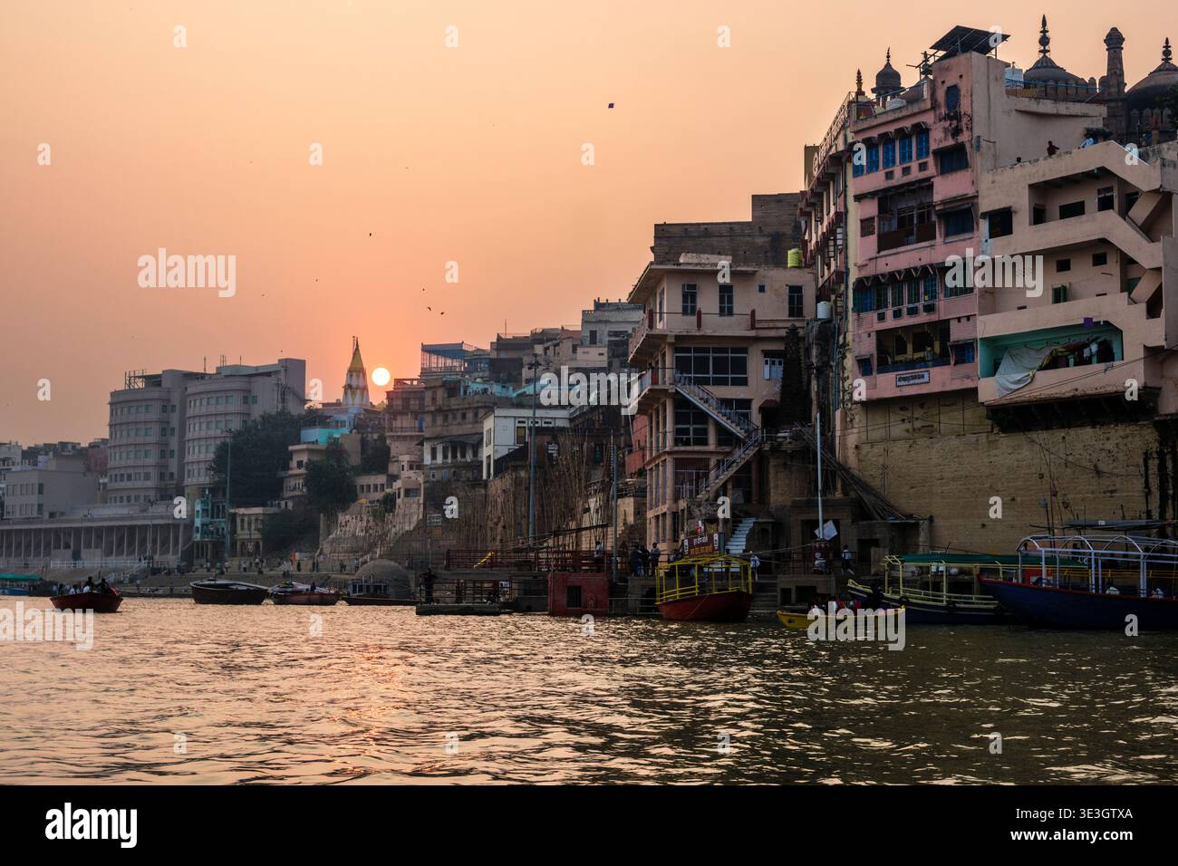 Il sole tramonta dietro i fitti edifici del lungofiume Varanasi mentre le barche si spostano attraverso il fiume Gange vicino ai ghat storici. Foto Stock