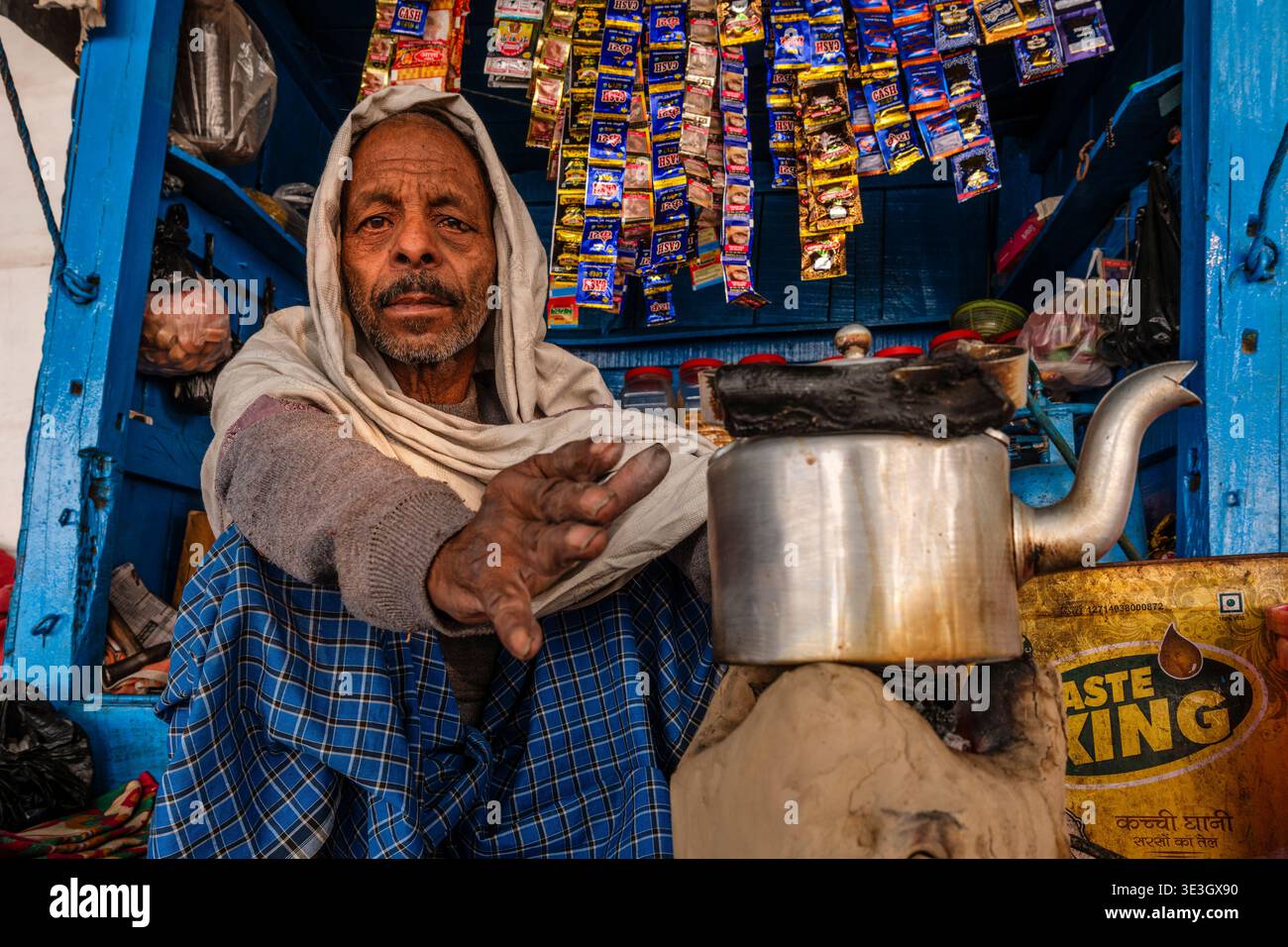 Un venditore ambulante a Varanasi siede all'interno del suo piccolo chiosco di legno blu mentre prepara il tè in un grande bollitore di metallo. Foto Stock