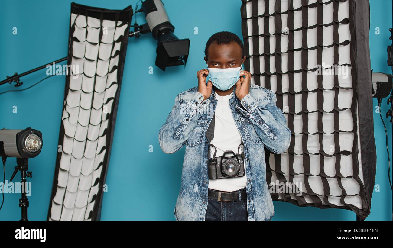 Modello maschile in posa con attrezzature per il backstage fotografico durante la pandemia del COVID 19. Fotografo con cavalletto, proiettori, fotocamera reflex digitale e obiettivo professionale nello studio dietro le quinte. Foto Stock
