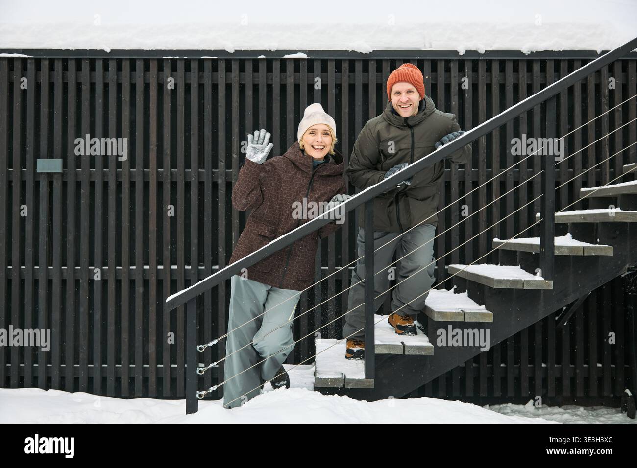 Donna caucasica di mezza età che sventola e sorride mentre sta in piedi su una scala innevata all'aperto accanto a un uomo caucasico di mezza età sorridente, entrambi indossano cappelli e guanti invernali in ambiente invernale Foto Stock