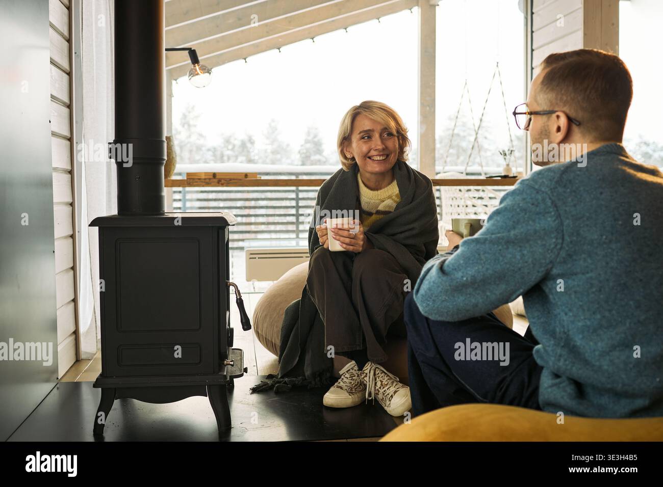 Donna caucasica di mezza età sorride e tiene in mano la tazza mentre siede vicino a una stufa a legna, parla con un uomo caucasico di mezza età, entrambe rilassanti all'interno in un'accogliente cabina Foto Stock