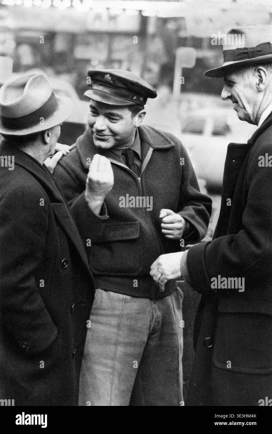 Questa accattivante fotografia di strada in bianco e nero degli anni '1940, catturata da Angelo Rizzuto, mostra tre uomini impegnati in una conversazione animata in quella che sembra essere Times Square, New York City. La figura centrale, che indossa un caratteristico cappello e una giacca, agisce energicamente parlando con i suoi compagni, che sono rivestiti di fedoras e cappotti, riflettendo la moda quotidiana dell'epoca. Foto Stock