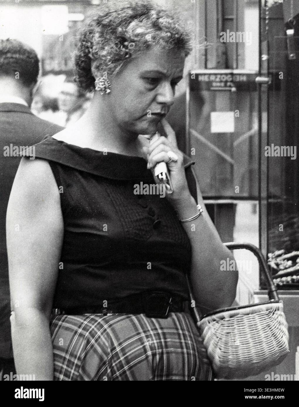 Questa accattivante fotografia di strada di Angelo Rizzuto, probabilmente degli anni '1950, cattura una donna di New York con un'espressione premurosa, tenendo in mano un accendisigari. È vestita in modo appropriato al periodo, con un top senza maniche scuro con un ampio colletto e una gonna tartan o a quadri, che riflette lo stile quotidiano della vita urbana della metà del secolo. Foto Stock