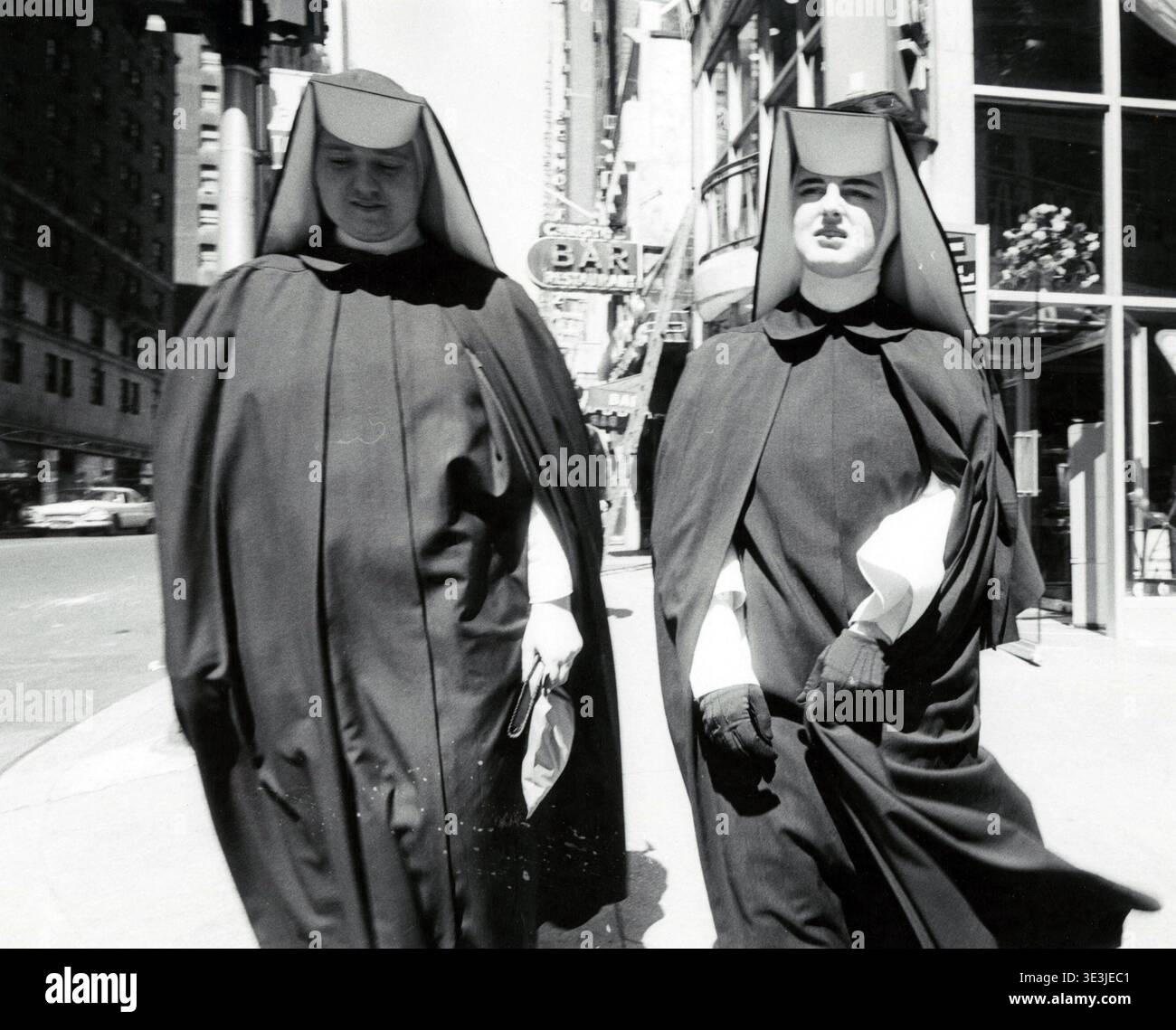 Questa accattivante fotografia di strada, probabilmente catturata da Angelo Rizzuto negli anni '1950 o '1960, raffigura due suore in abitudini tradizionali che camminano lungo un vivace marciapiede di New York. Gli stili contrastanti del loro abbigliamento sullo sfondo urbano offrono uno sguardo alla vita quotidiana e alla moda dell'epoca. Foto Stock