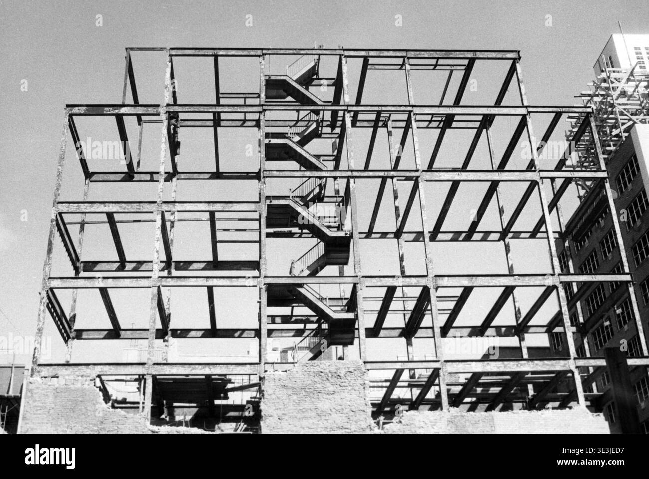 Questa fotografia in bianco e nero di Angelo Rizzuto degli anni '1940 cattura la struttura scheletrica in acciaio di un edificio in costruzione, probabilmente a New York. I motivi geometrici delle travi e una scalinata temporanea si distinguono contro il cielo limpido, mostrando un momento di sviluppo urbano. Foto Stock